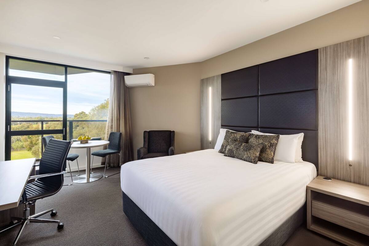 A hotel room with a large bed, a desk with two chairs, a small table, and a view of the outdoors.