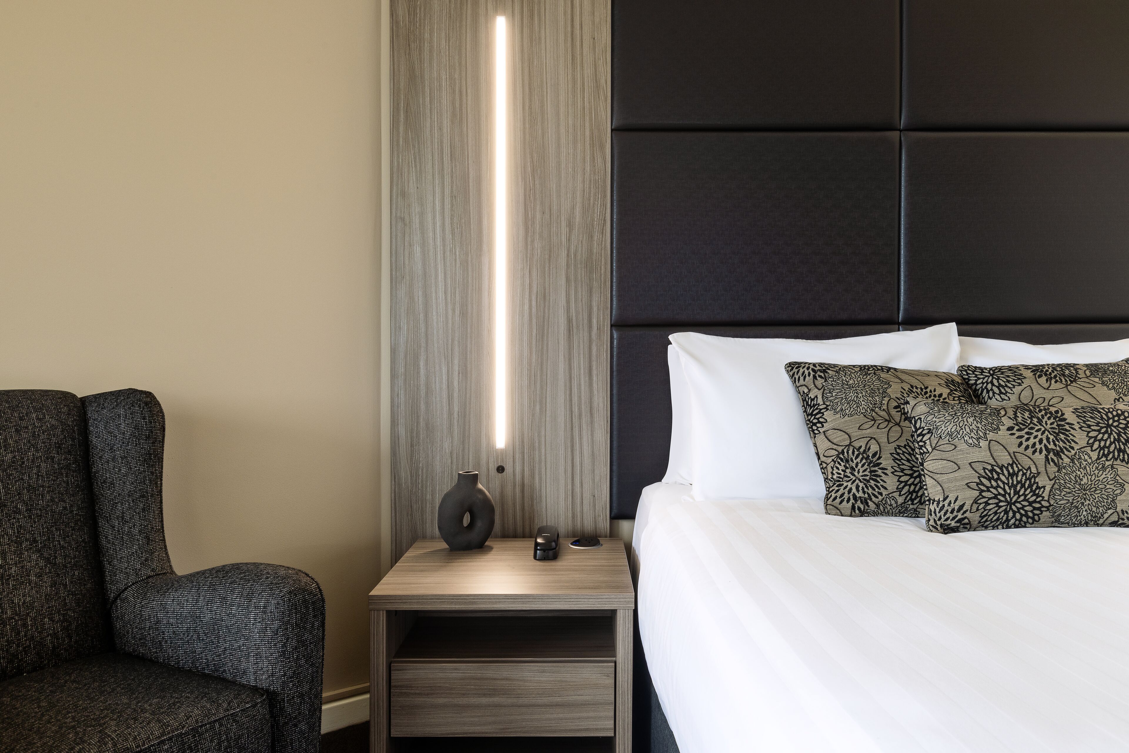 Ett modernt hotellrum med en säng, ett nattduksbord, en lampa och en stol.