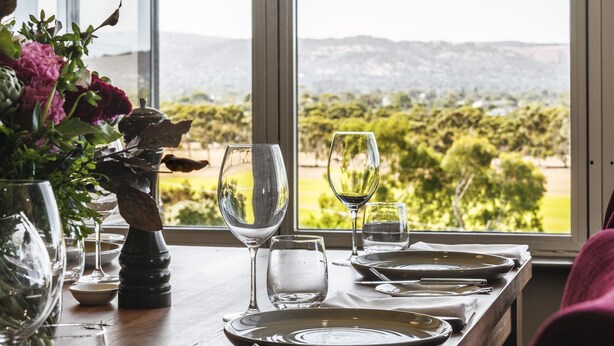 A dining table set with plates, glasses, and a vase of flowers, with a scenic view through a window. - Rydges South Park Adelaide (Adelaide)