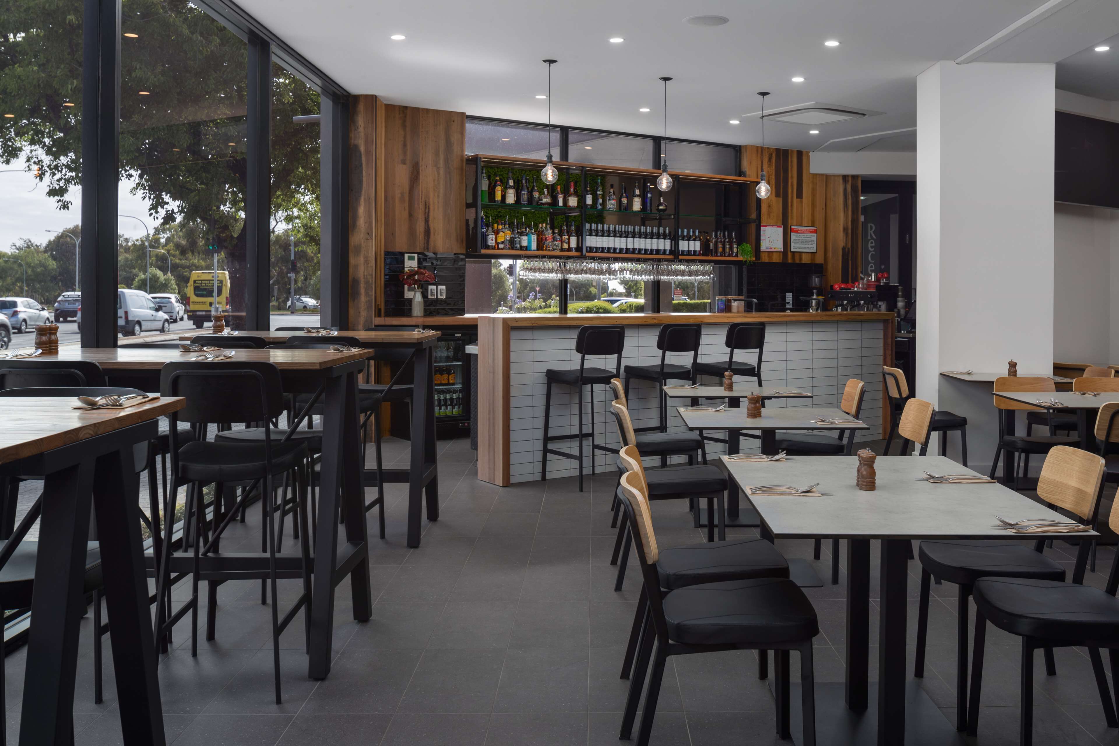A modern restaurant with a bar area, multiple tables and chairs, and a view of the street through large windows.