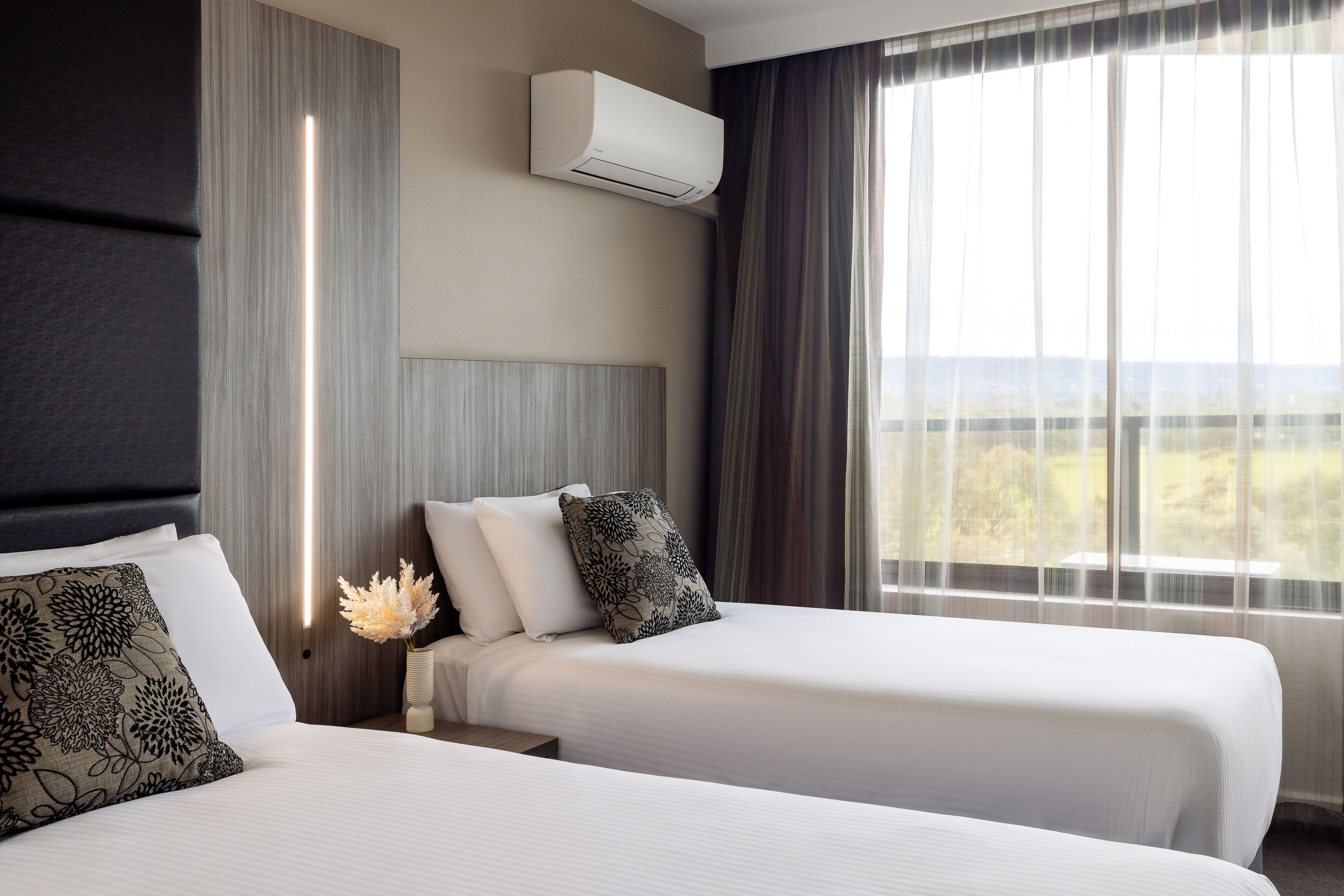 A hotel room with two beds, a large window with curtains, an air conditioning unit, and a nightstand with a vase of flowers.
