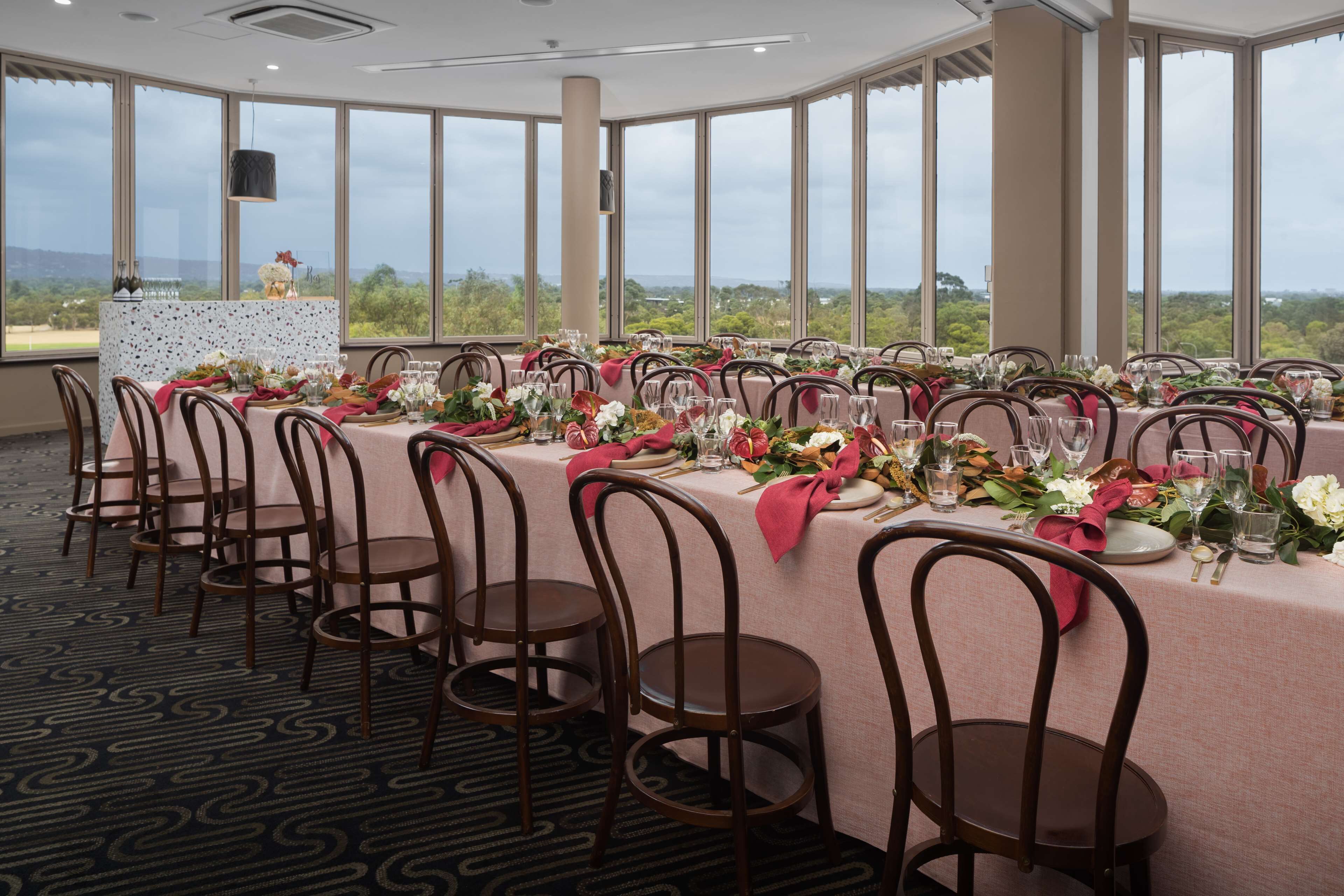 A banquet hall with a long table set for a formal event, surrounded by chairs, and large windows offering a view of the outdoors.