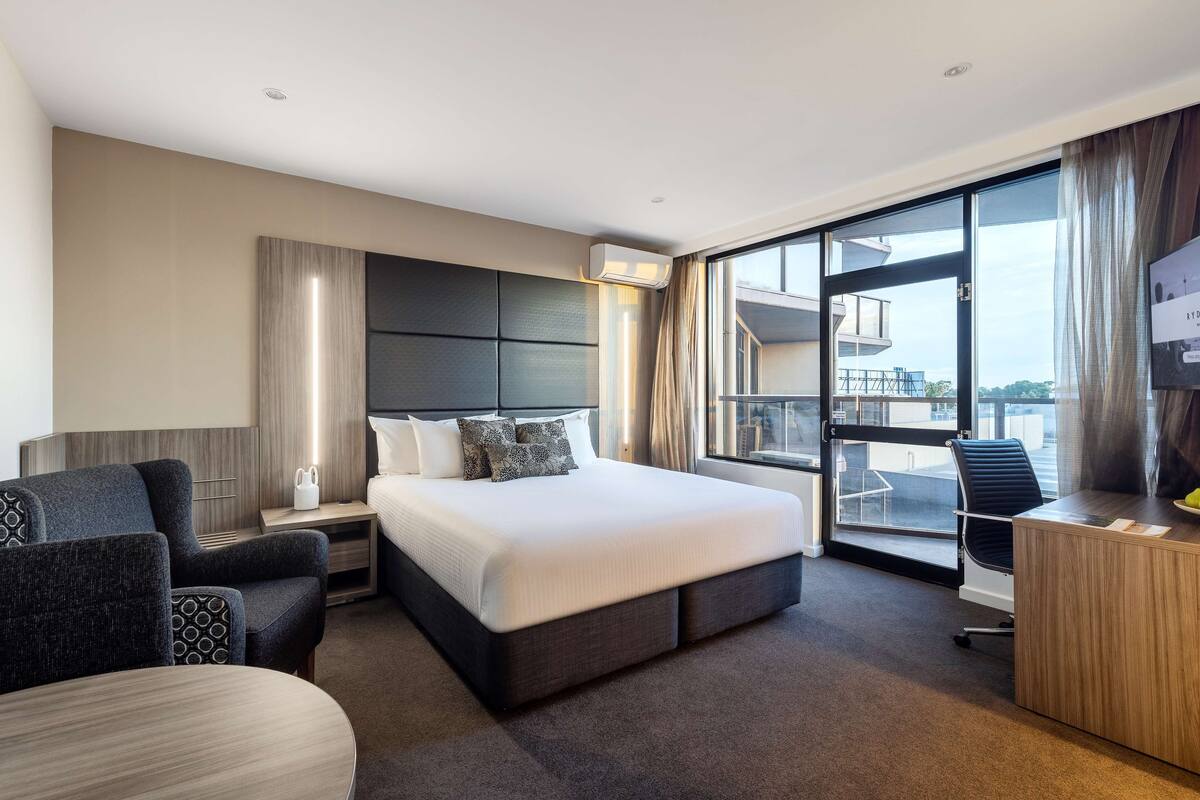 A modern hotel room with a large bed, a desk, and a view of the city through large windows.