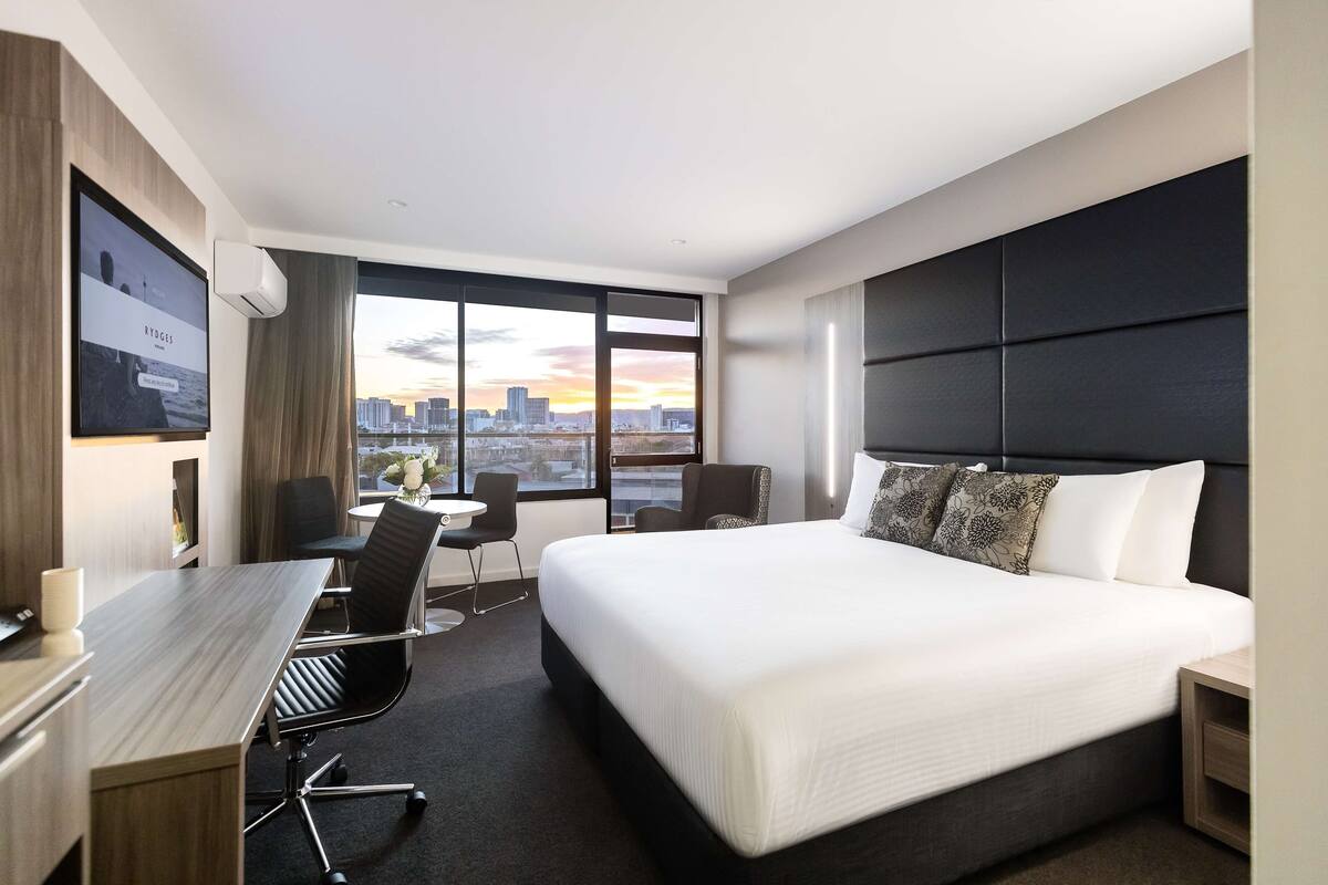 A hotel room with a large bed, a desk with a chair, a small table, and a large window with a city view.
