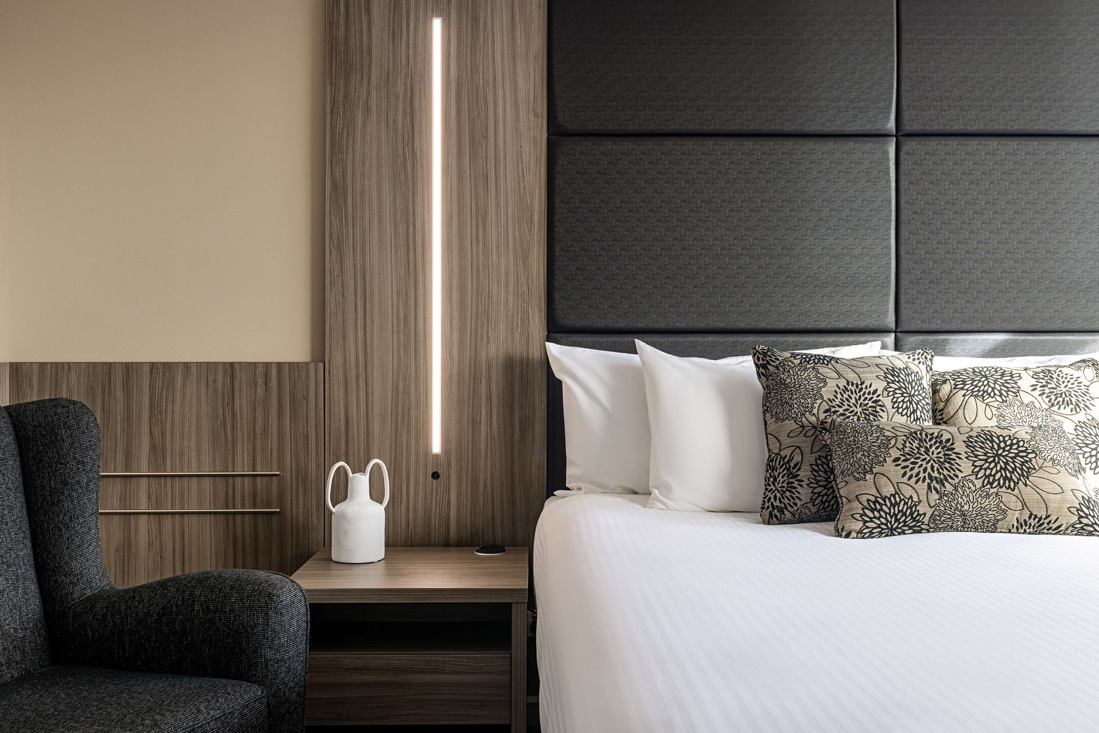 Une chambre d’hôtel moderne avec une tête de lit en bois, une table de chevet dotée d’une lampe blanche et un lit agrémenté d’oreillers à motifs.
