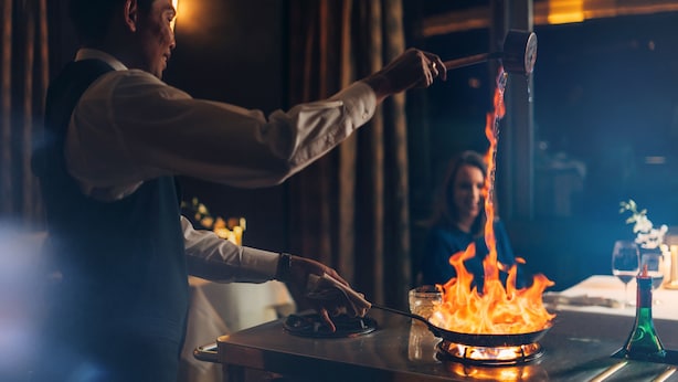 Une personne cuisine sur une cuisinière, utilisant des flammes, dans une pièce faiblement éclairée.