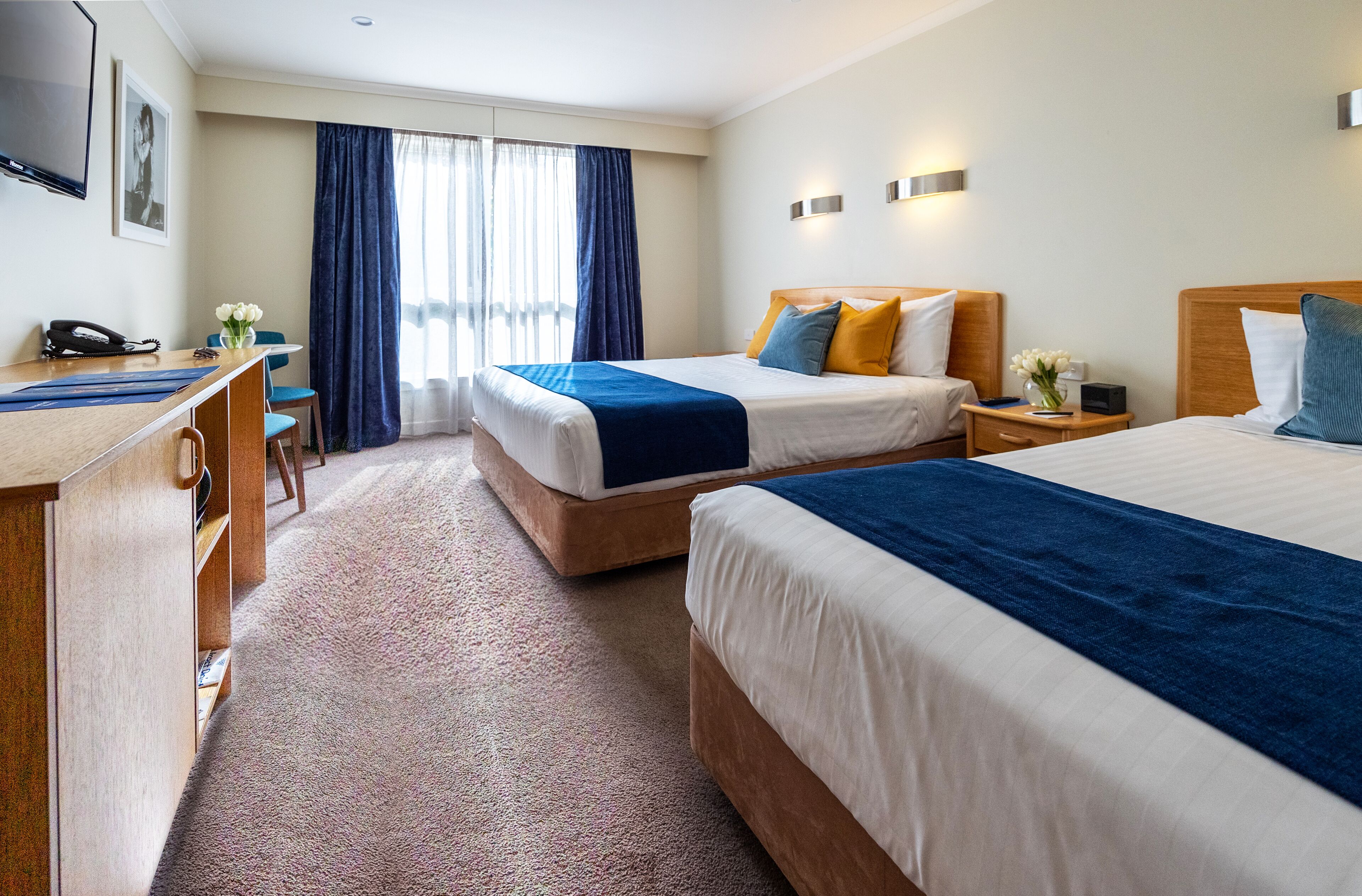Une chambre d’hôtel avec deux lits, un bureau, une chaise, un téléphone et une fenêtre avec des rideaux.