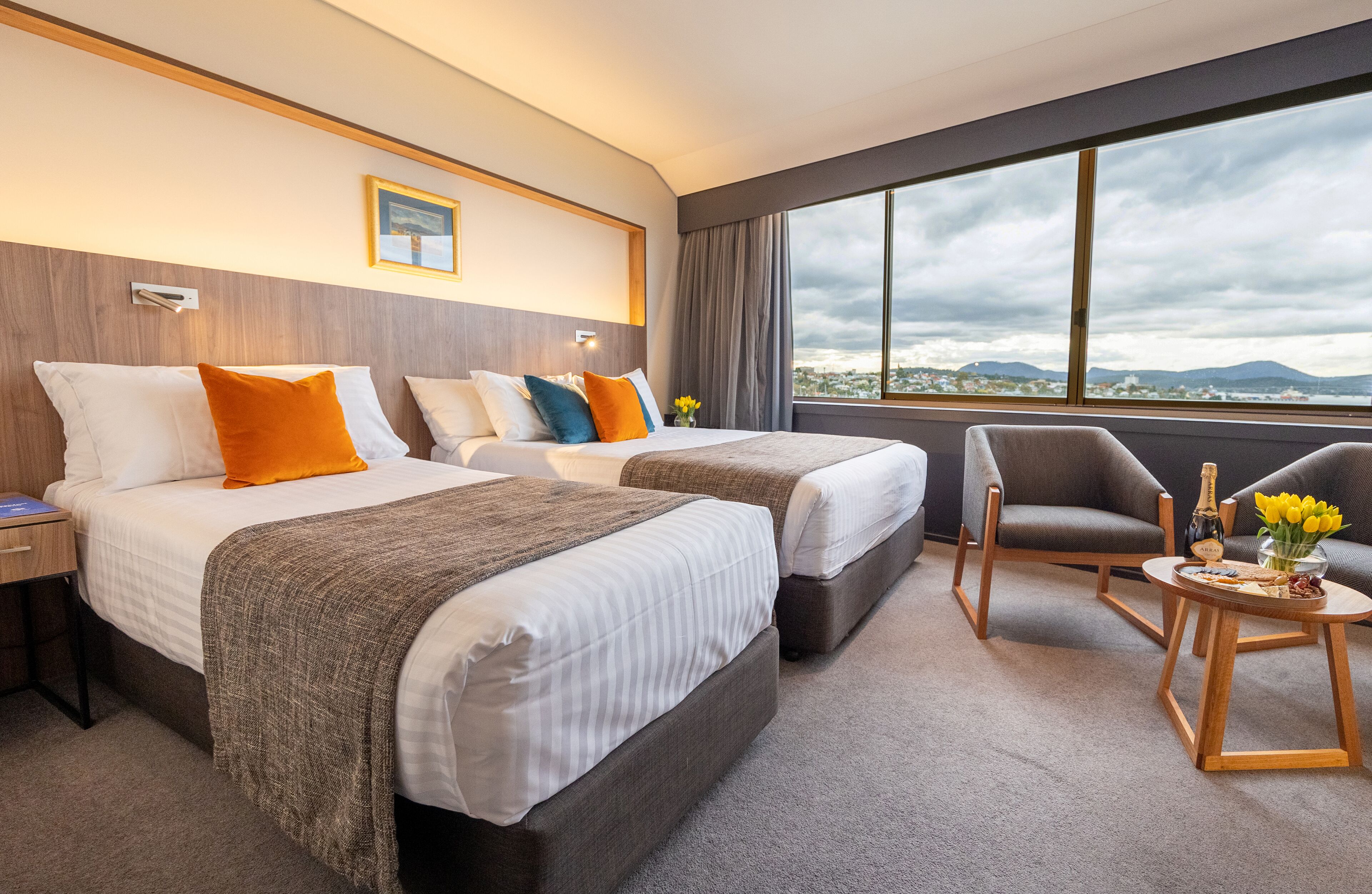 A hotel room with two beds, a chair, a small table, and a large window offering a city view.