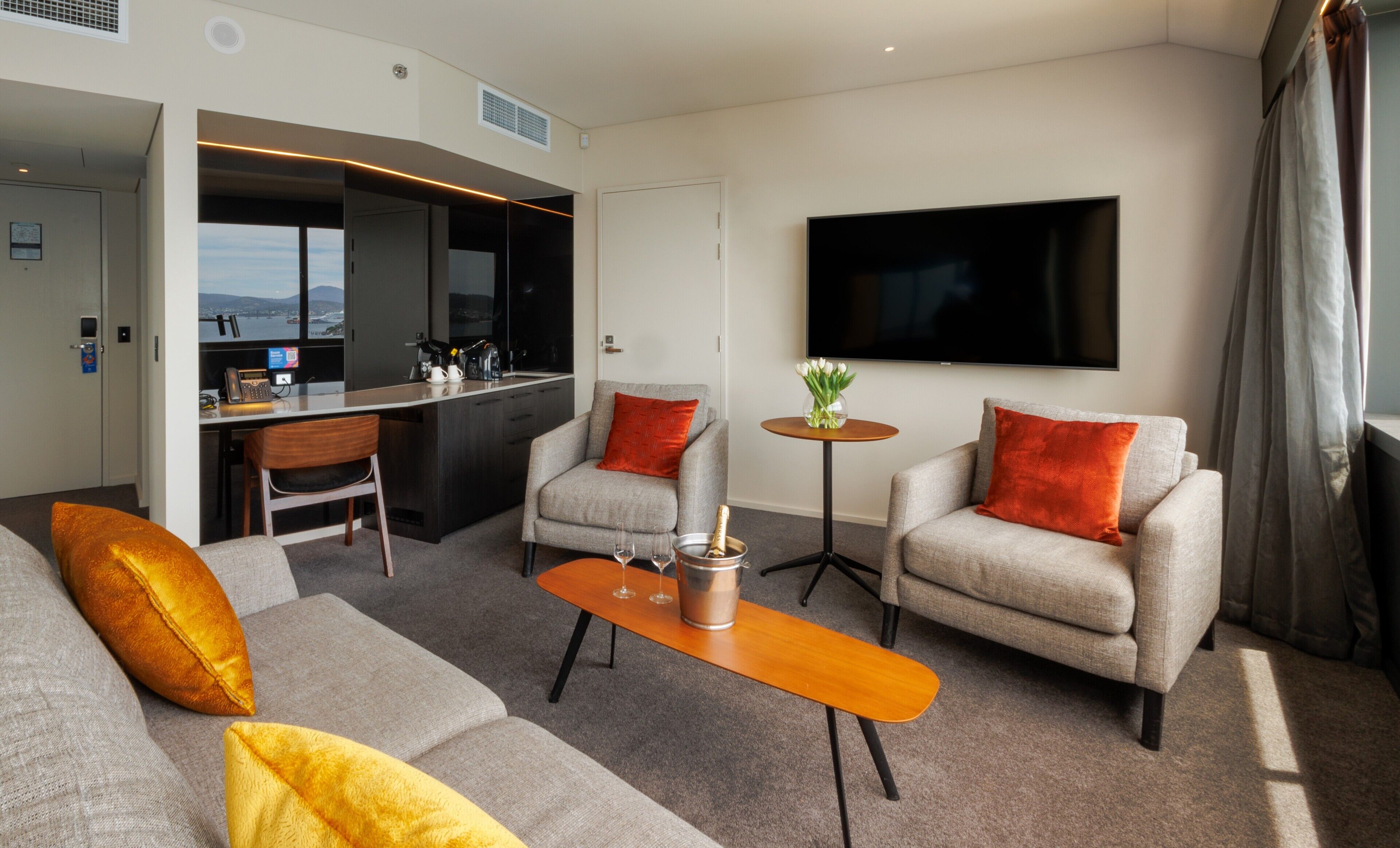 A modern hotel room with a sofa, armchairs, a coffee table, a TV, and a view of the city.