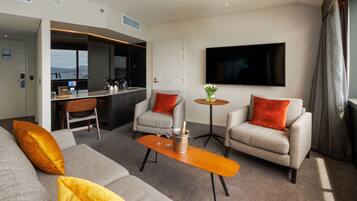 A modern hotel room with a sofa, armchairs, a coffee table, a TV, and a view of the city.