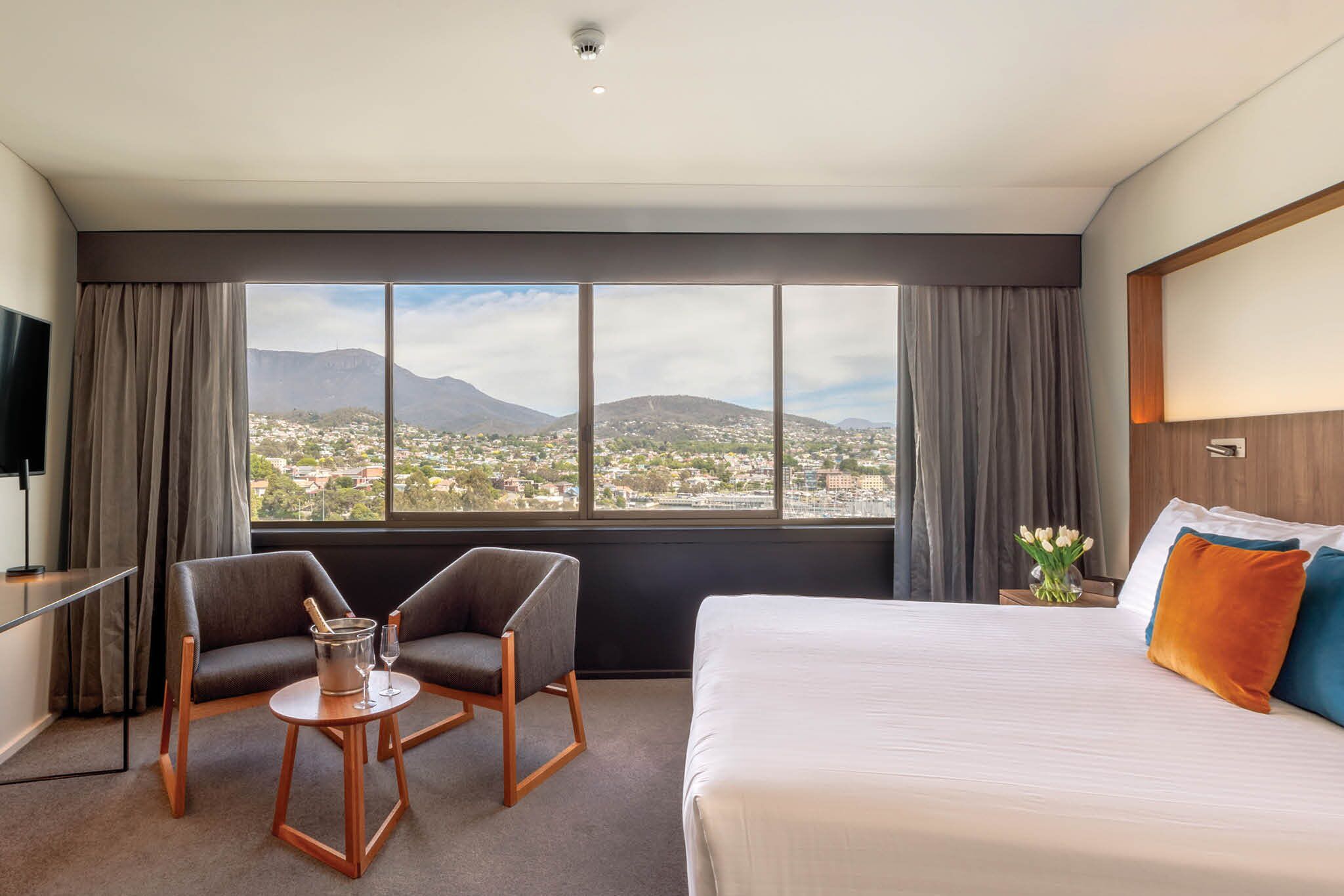 Habitación de hotel con un ventanal grande que ofrece vistas a la ciudad, una cama con dos almohadas, una silla y una mesita con una botella y un vaso.