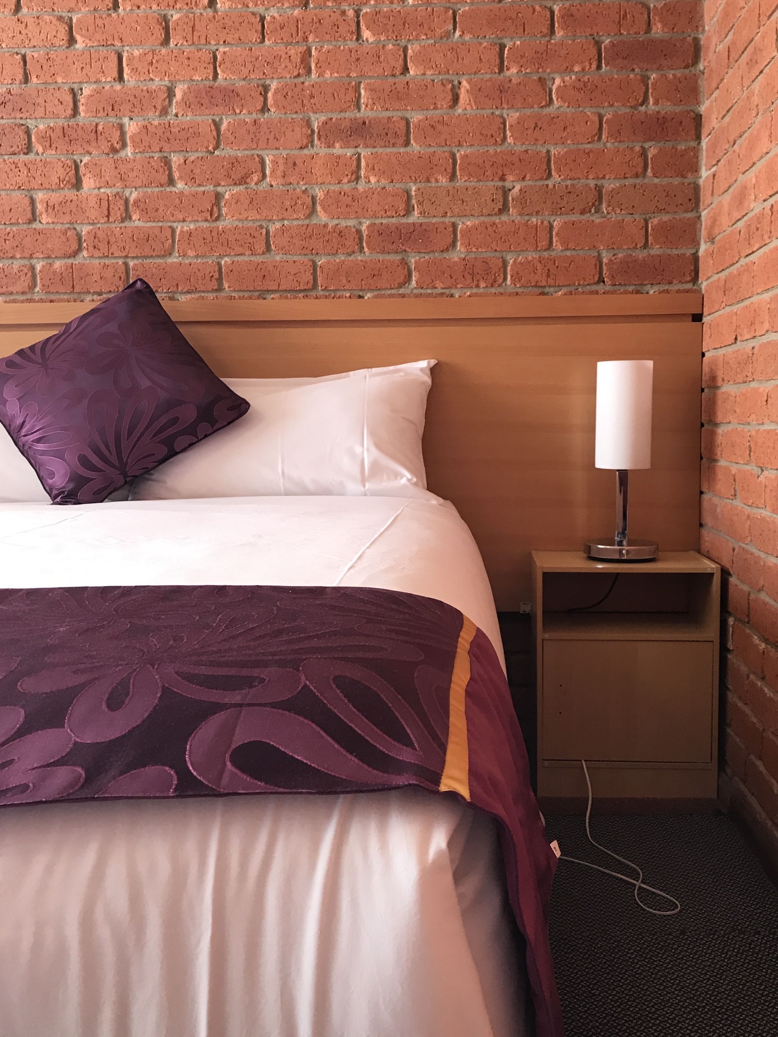 Habitación de hotel con pared de ladrillo, cabecera de madera, cama con ropa de cama blanca y morada, y mesita de noche con lámpara.