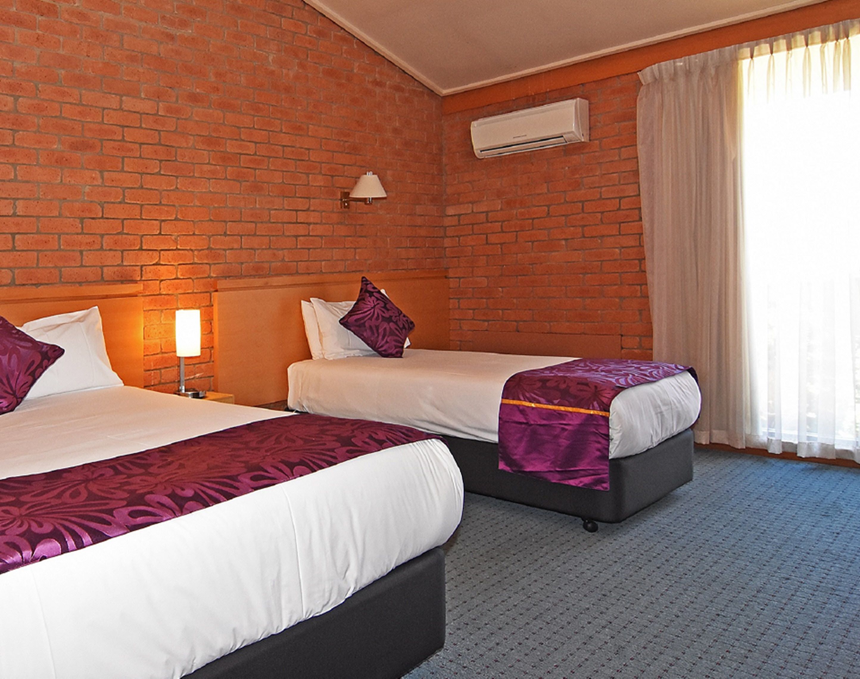 Habitación de hotel con dos camas, una pared de ladrillo, un equipo de aire acondicionado y una ventana con cortinas.