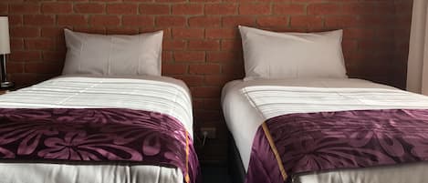 Two beds with white linens and purple floral bedspreads against a brick wall.