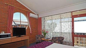 A hotel room with a brick wall, a television on a wooden dresser, a bed with a purple bedspread, a small table with chairs, and a window with lace curtains.