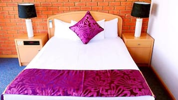 A hotel room with a brick wall, a bed with white bedding and a purple floral bedspread, two bedside tables with lamps, and a digital clock on one of the tables.