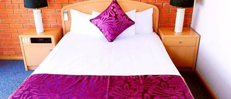 A hotel room with a brick wall, a bed with white bedding and a purple floral bedspread, two bedside tables with lamps, and a digital clock on one of the tables.