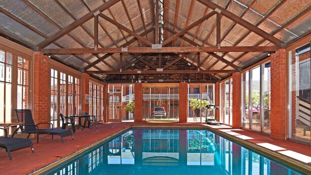 An indoor swimming pool with a high ceiling and brick walls, surrounded by windows and seating areas.