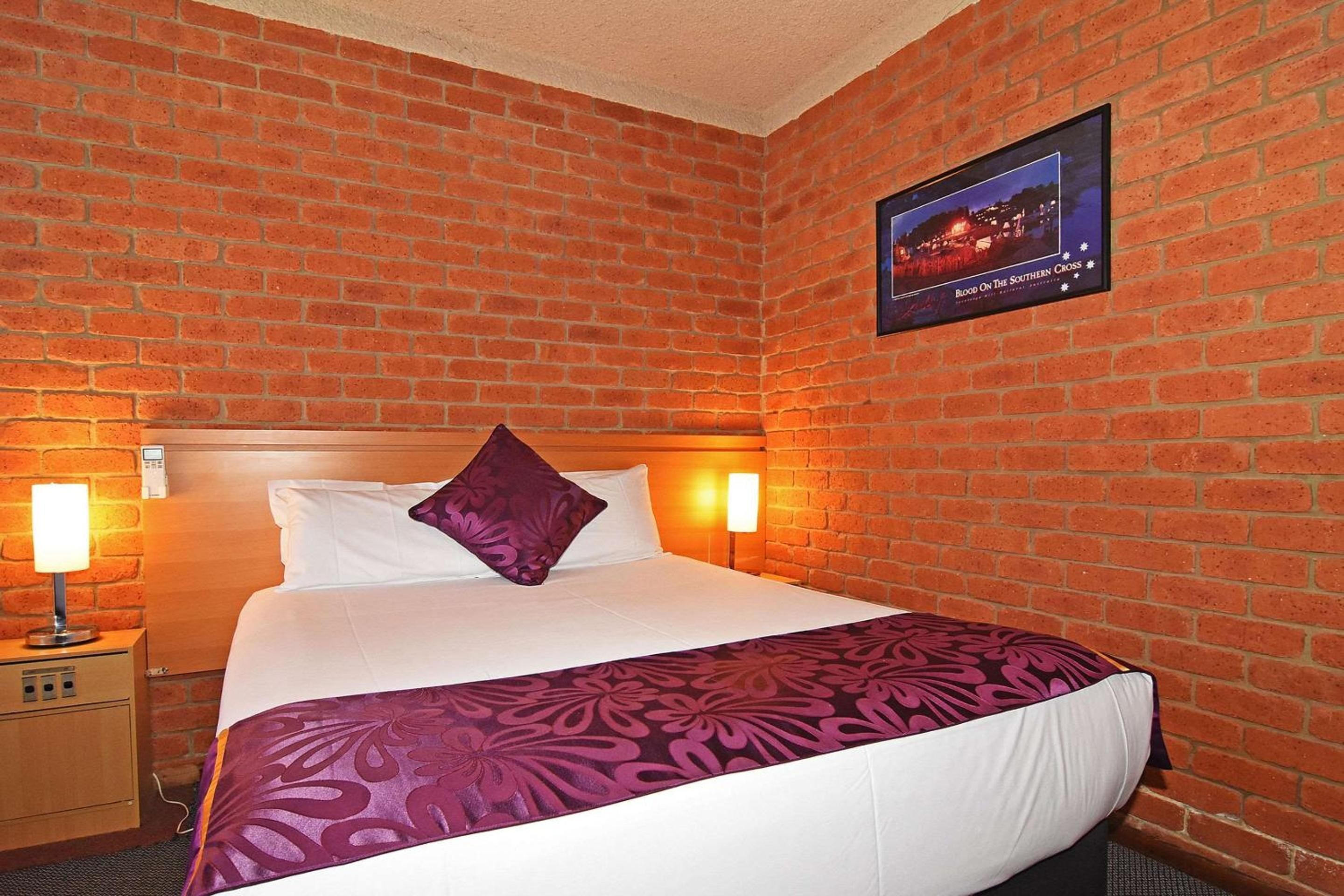 A hotel room with a brick wall, a bed with white linens and a purple throw, two bedside lamps, and a framed picture on the wall.