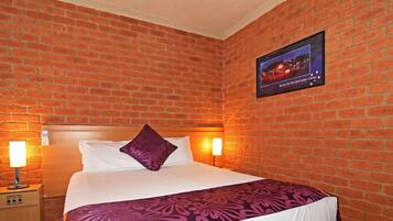 A hotel room with a brick wall, a bed with white linens and a purple throw, two bedside lamps, and a framed picture on the wall.