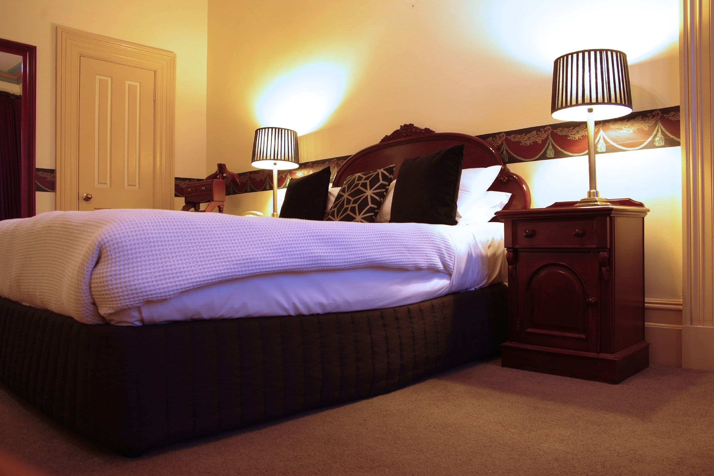 A neatly made bed with a patterned bedspread and pillows, a wooden nightstand with a lamp, and a door in the background.