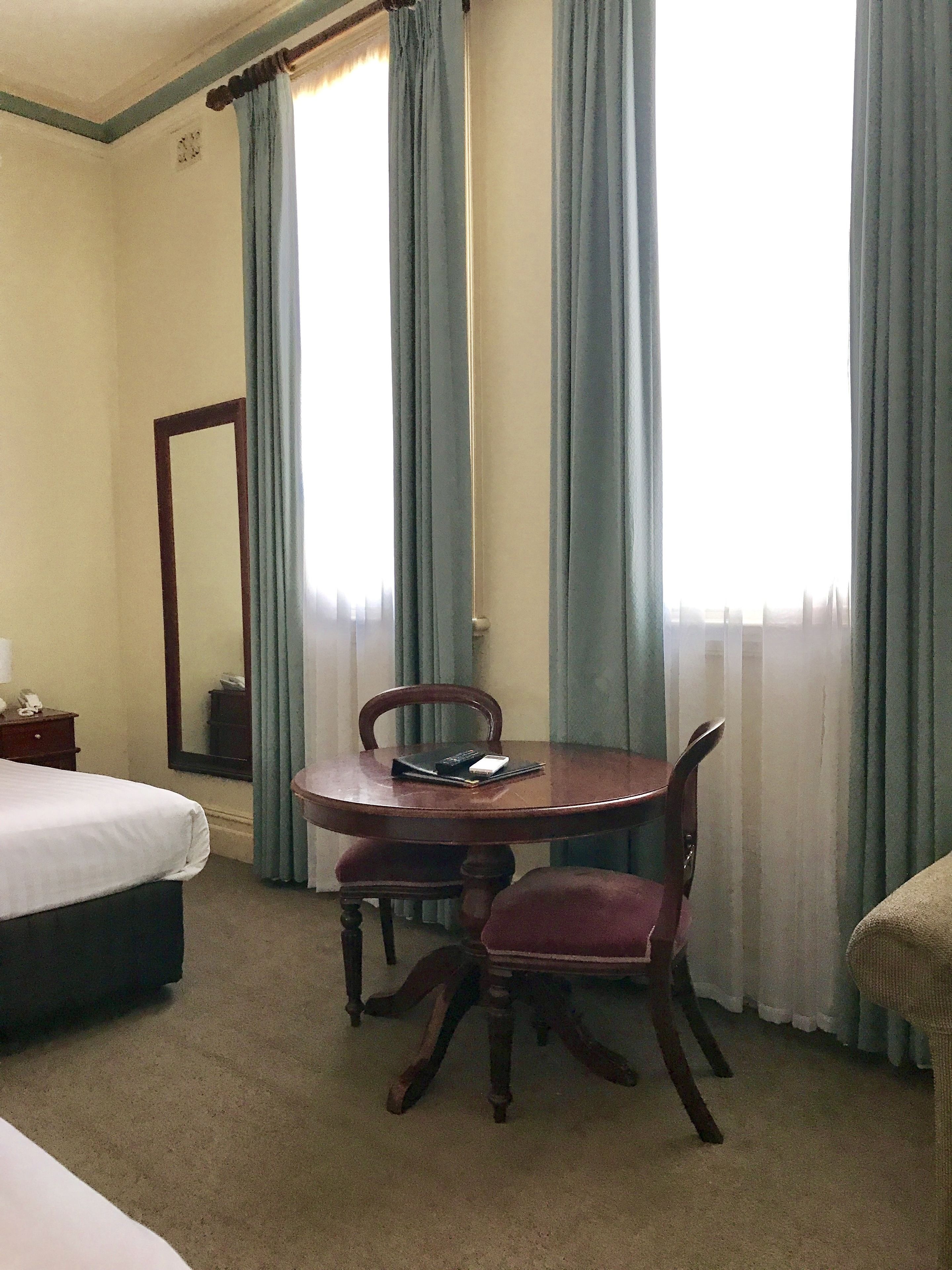 A hotel room with a bed, a wooden dining table with chairs, a mirror, and a window with curtains.