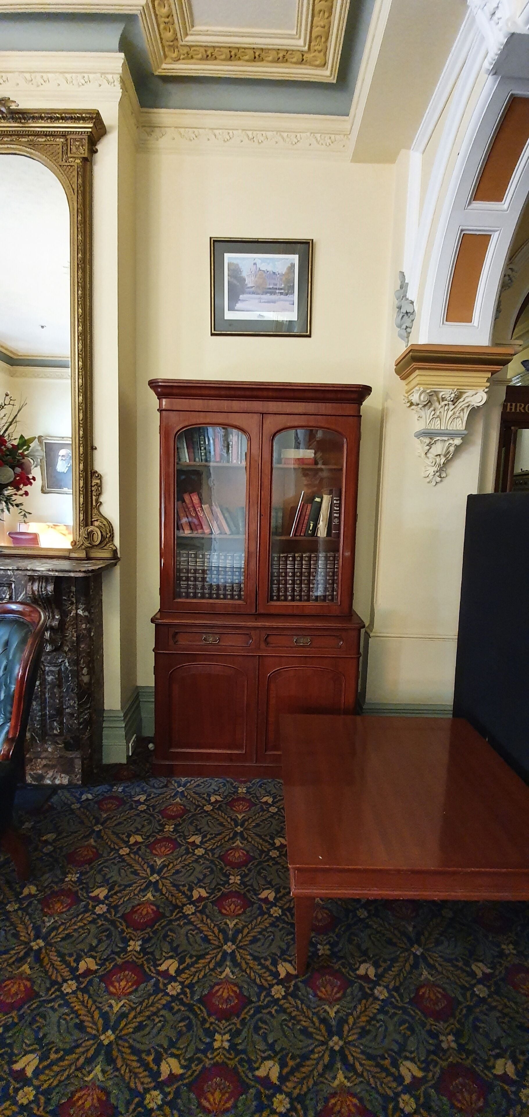 A room with a patterned carpet, a wooden cabinet with glass doors, a framed picture on the wall, and a large mirror.