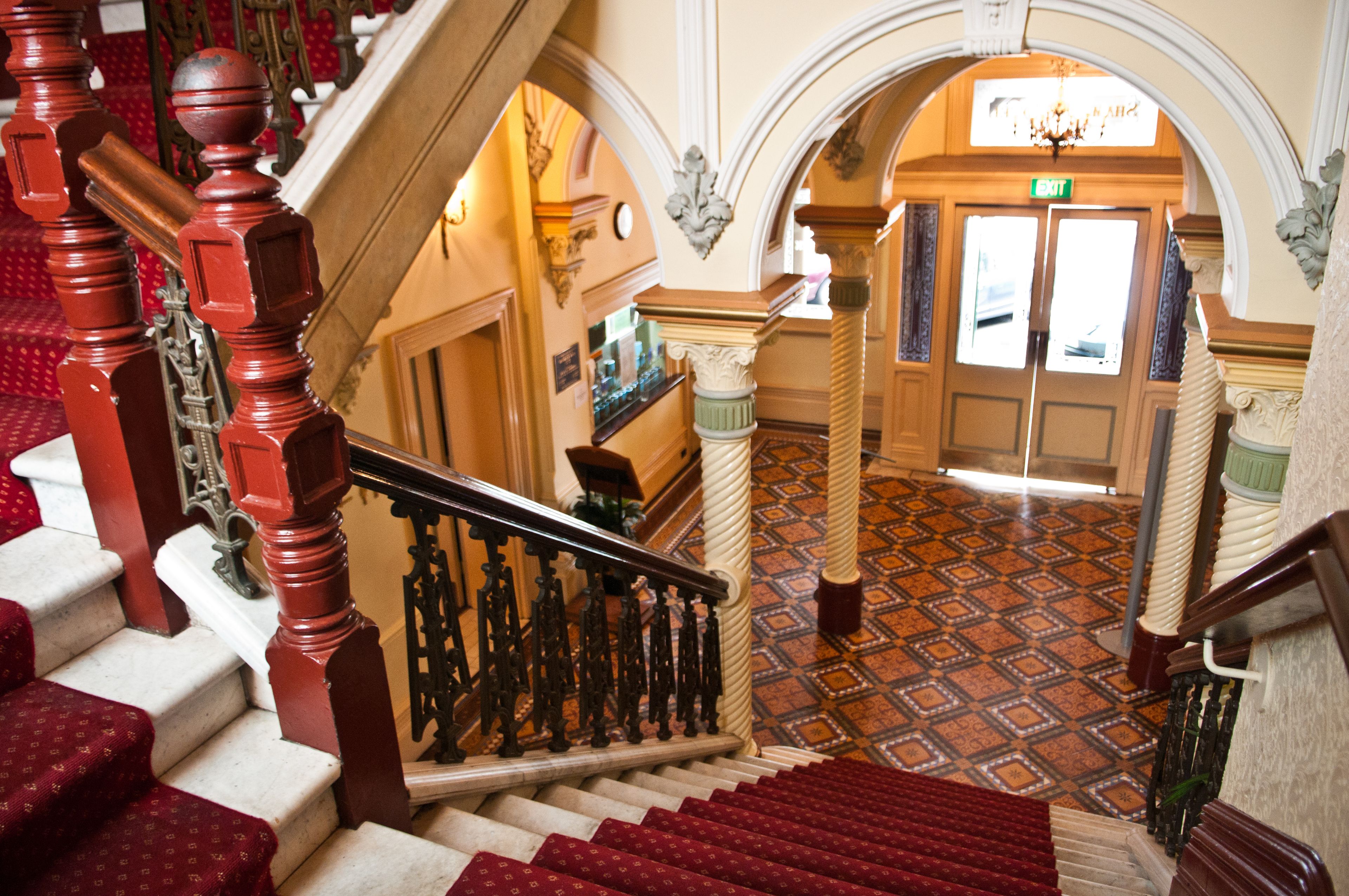A grand staircase with ornate railings and red carpeted steps leading to a well-lit hallway with patterned flooring and decorative columns.
