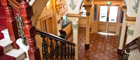 A grand staircase with ornate railings and red carpeted steps leading to a well-lit hallway with patterned flooring and decorative columns.