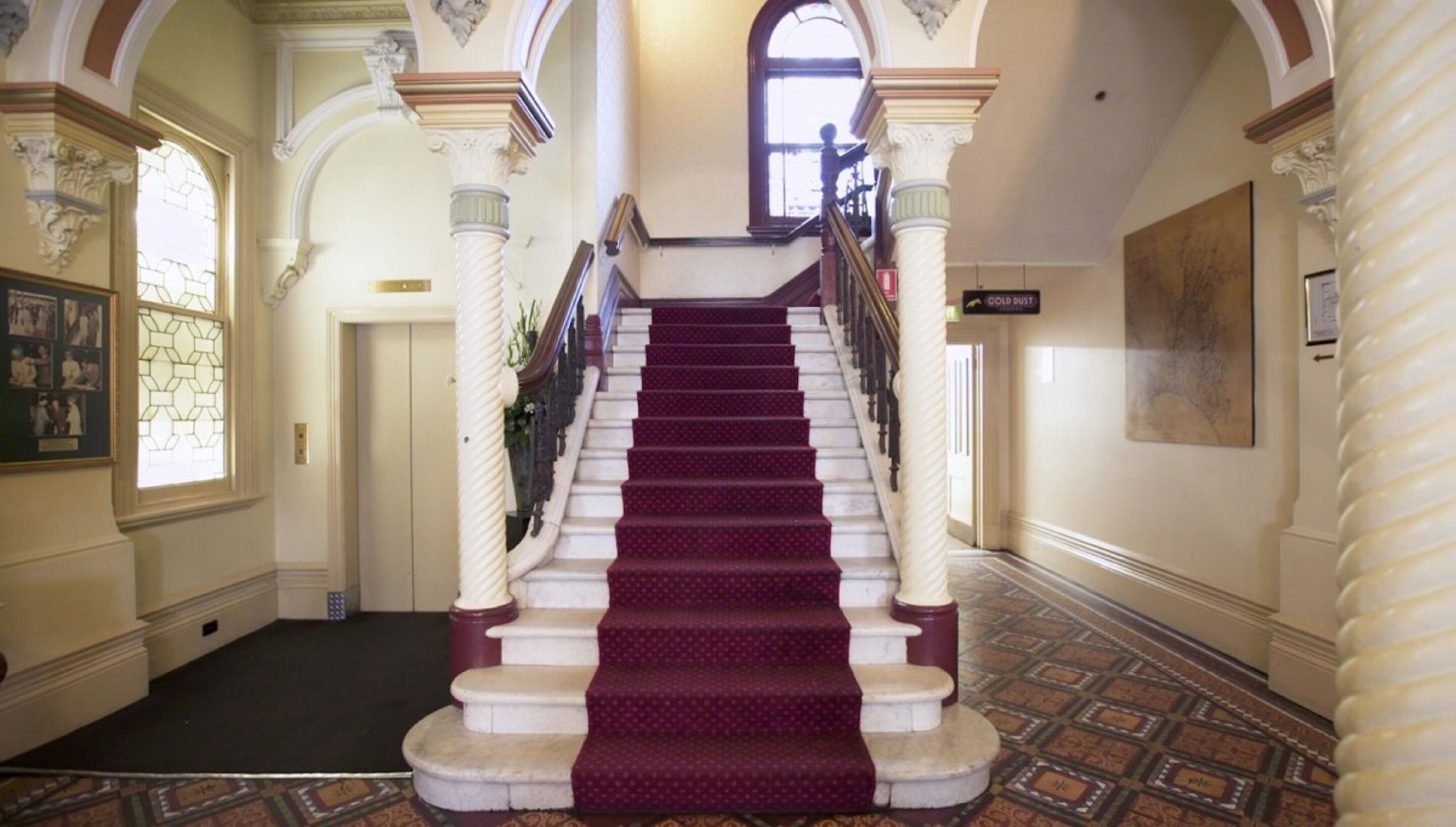 Un magnifique escalier recouvert d’un tapis rouge, flanqué de colonnes richement décorées et surmonté d’une grande fenêtre.