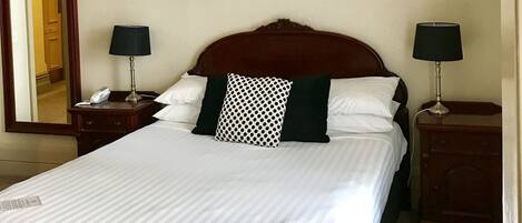 Un lit bien fait, avec une literie blanche et un coussin à motifs, flanqué de deux tables de chevet avec des lampes, une tête de lit en bois et un miroir au mur.