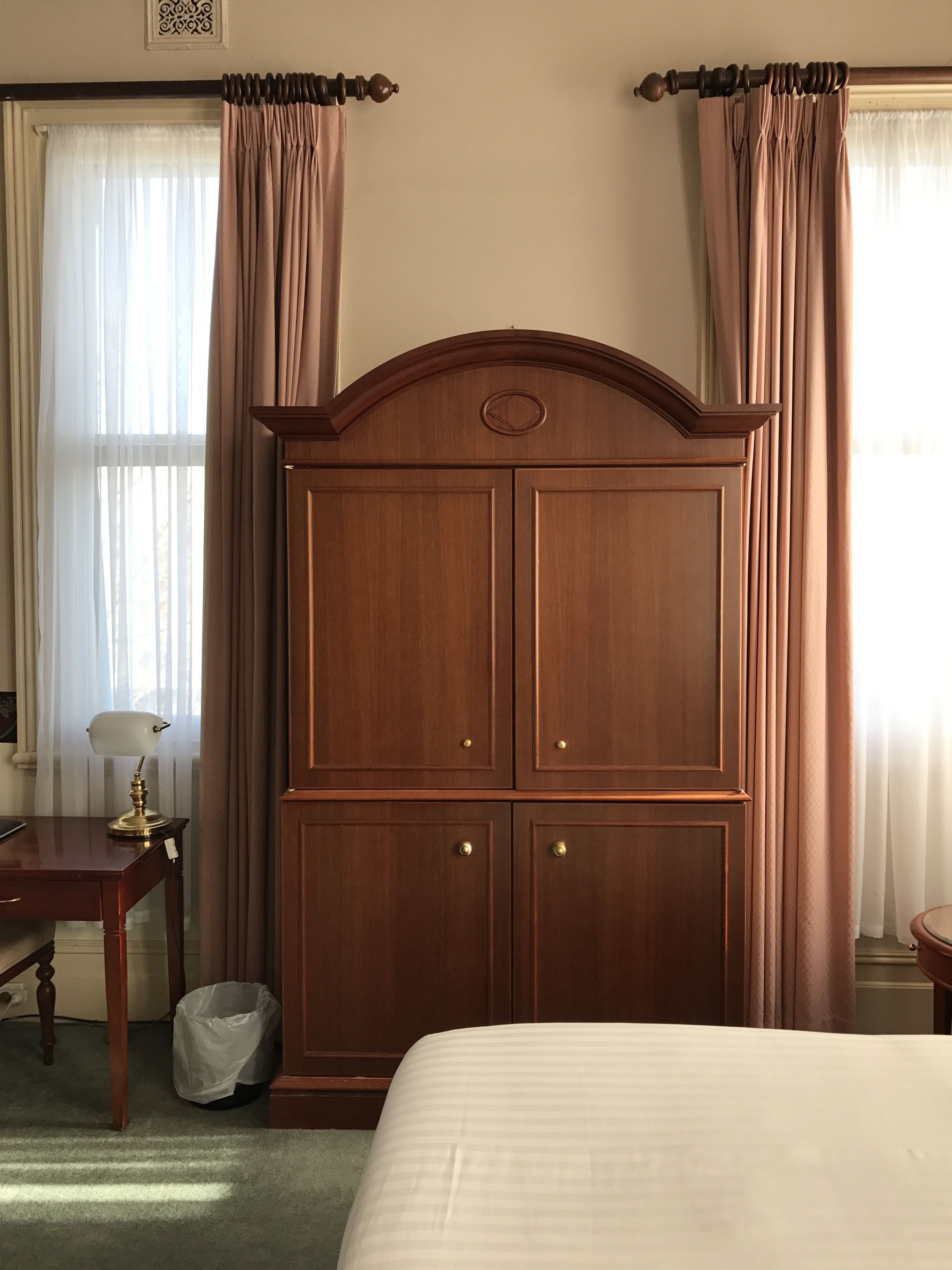 A bedroom with a large wooden wardrobe, a bed, a desk with a lamp, and sheer curtains.