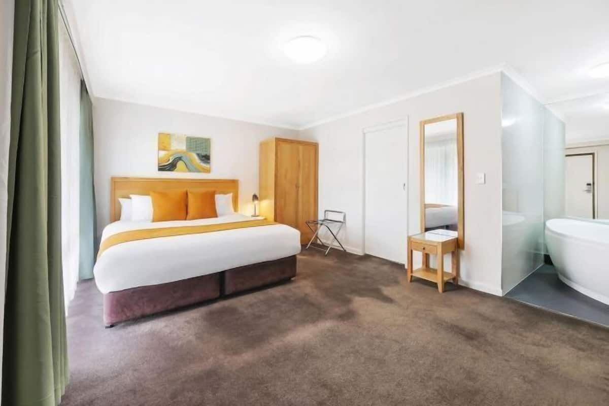 A hotel room with a bed, a small table, a mirror, and a bathroom with a bathtub.