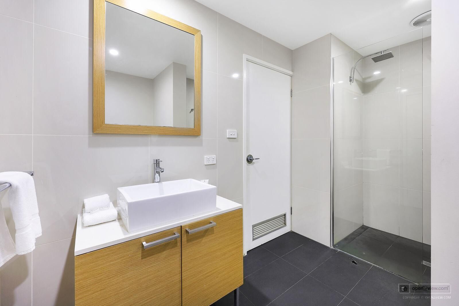 Un bagno moderno con lavandino bianco, mobile contenitore in legno, uno specchio grande e box doccia in vetro.