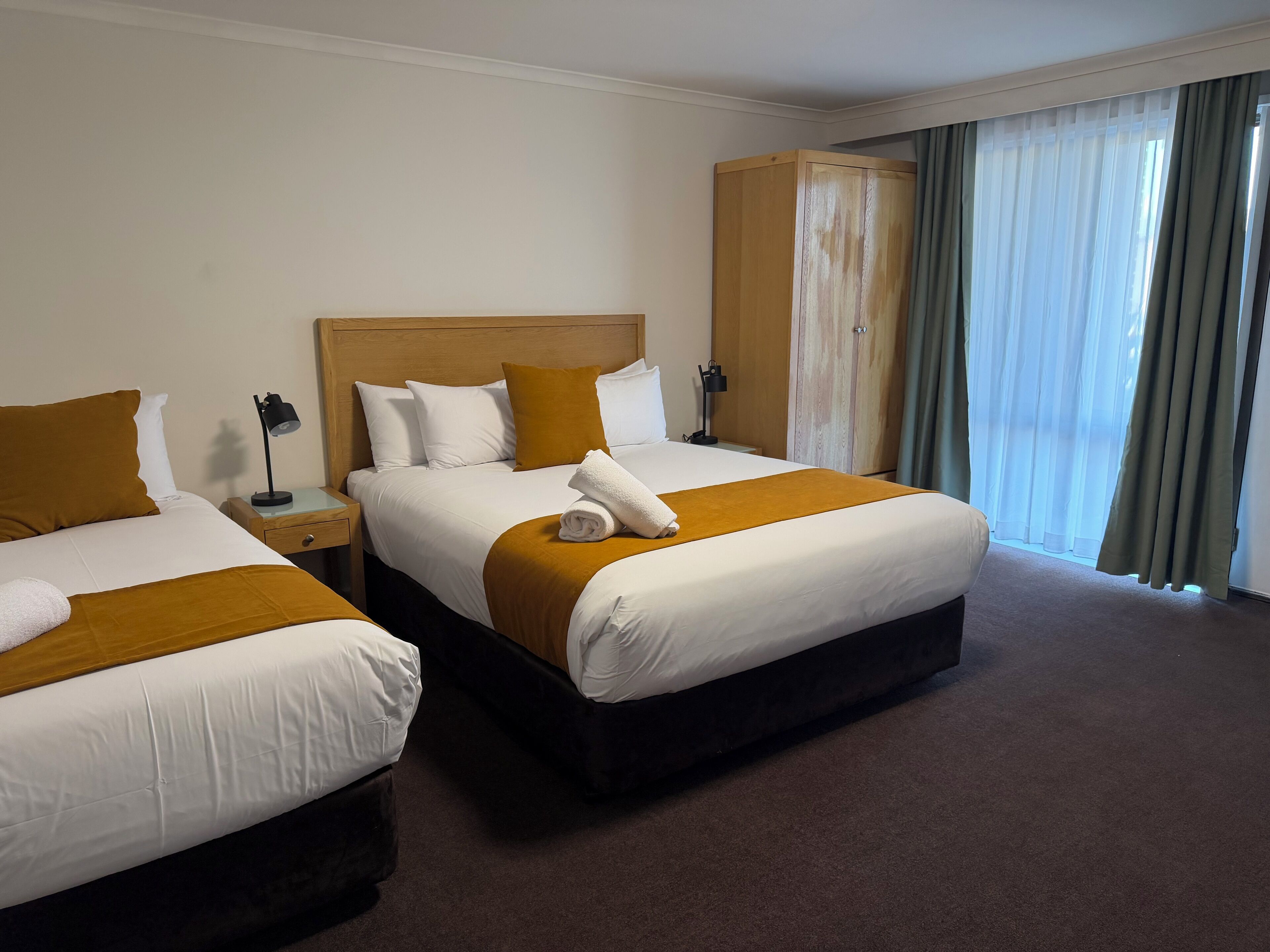 A hotel room with two beds, a wooden headboard, a nightstand with a lamp, and a large window with curtains.