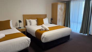 A hotel room with two beds, a wooden headboard, a nightstand with a lamp, and a large window with curtains.