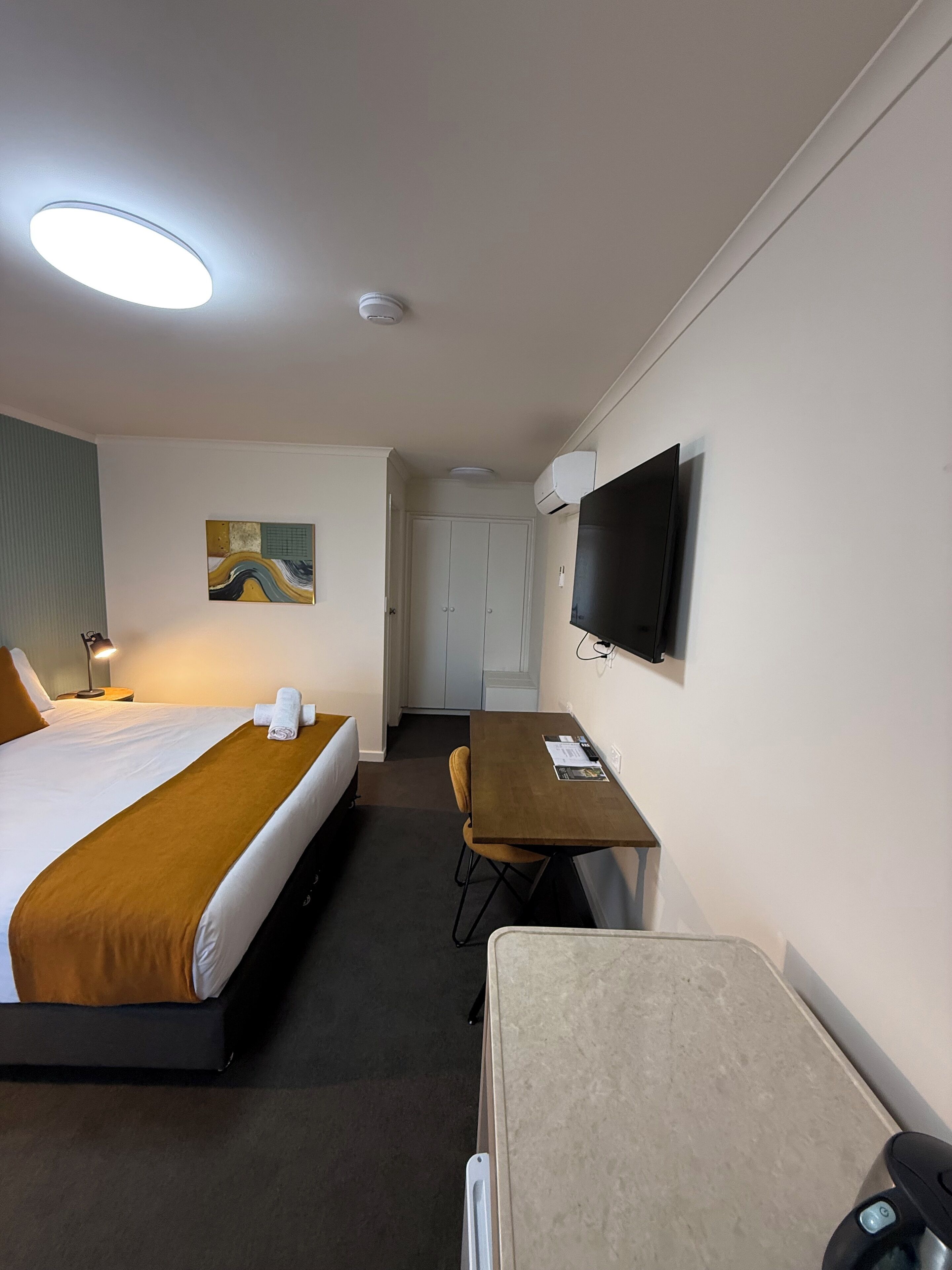 A hotel room with a bed, a desk, a chair, a TV, and an air conditioning unit.