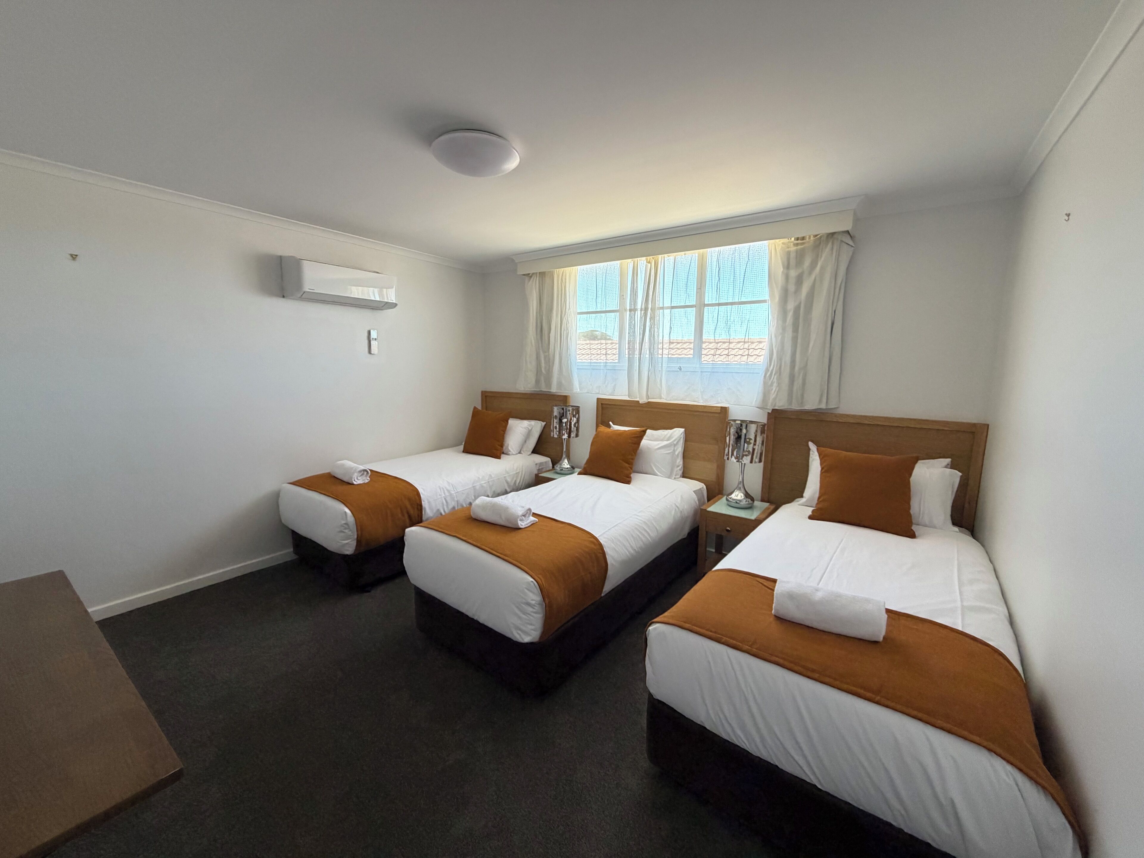 Una habitación de hotel con tres camas individuales, una mesita de noche, una lámpara y una ventana con cortinas.