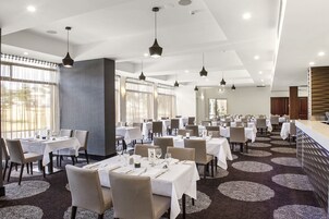 Un comedor amplio con varias mesas y sillas, ventanas grandes con cortinas e iluminación colgante.