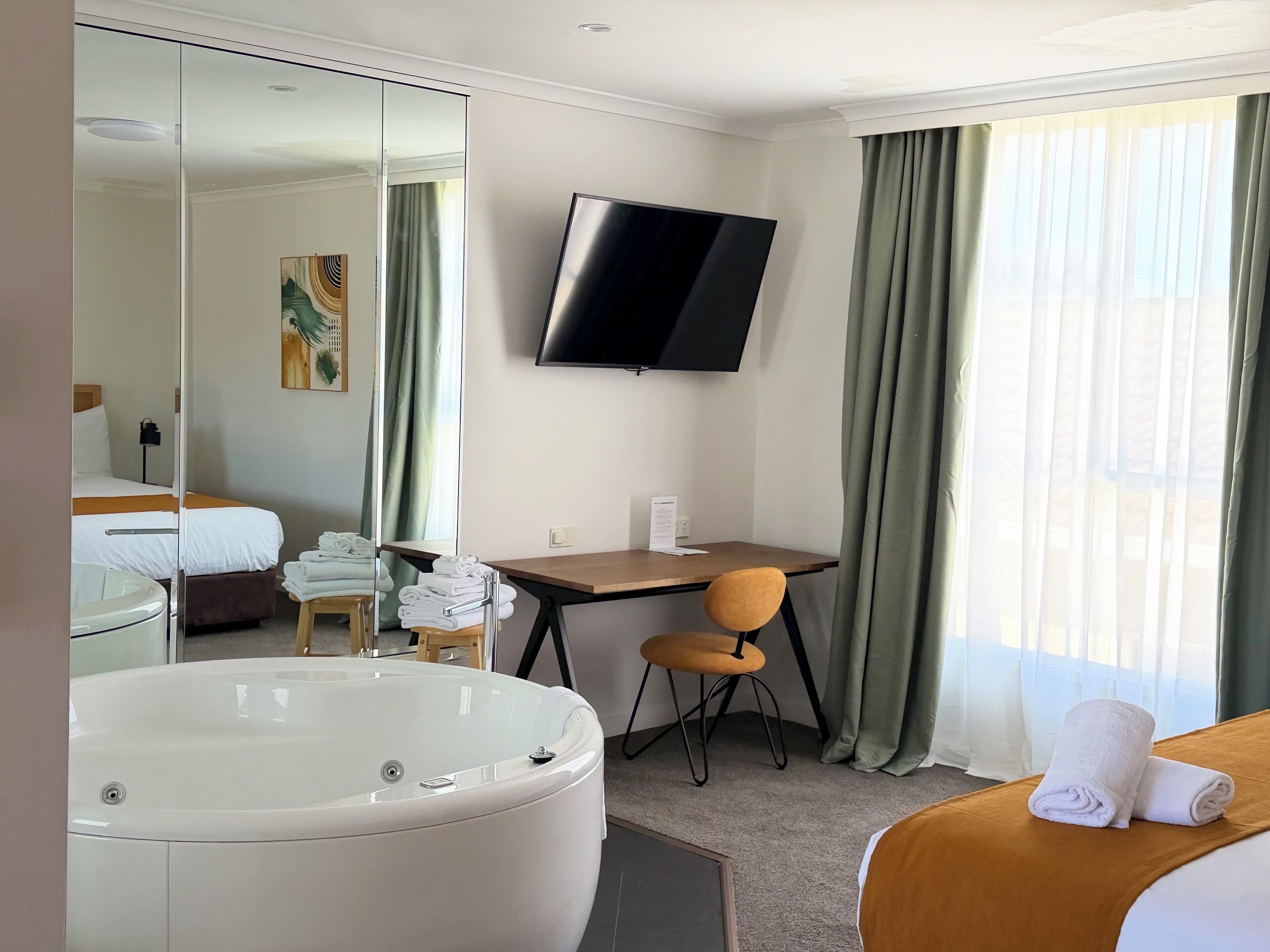 A hotel room with a large bathtub, a bed with a brown headboard, a desk with a chair, a flat-screen TV mounted on the wall, and a window with curtains.