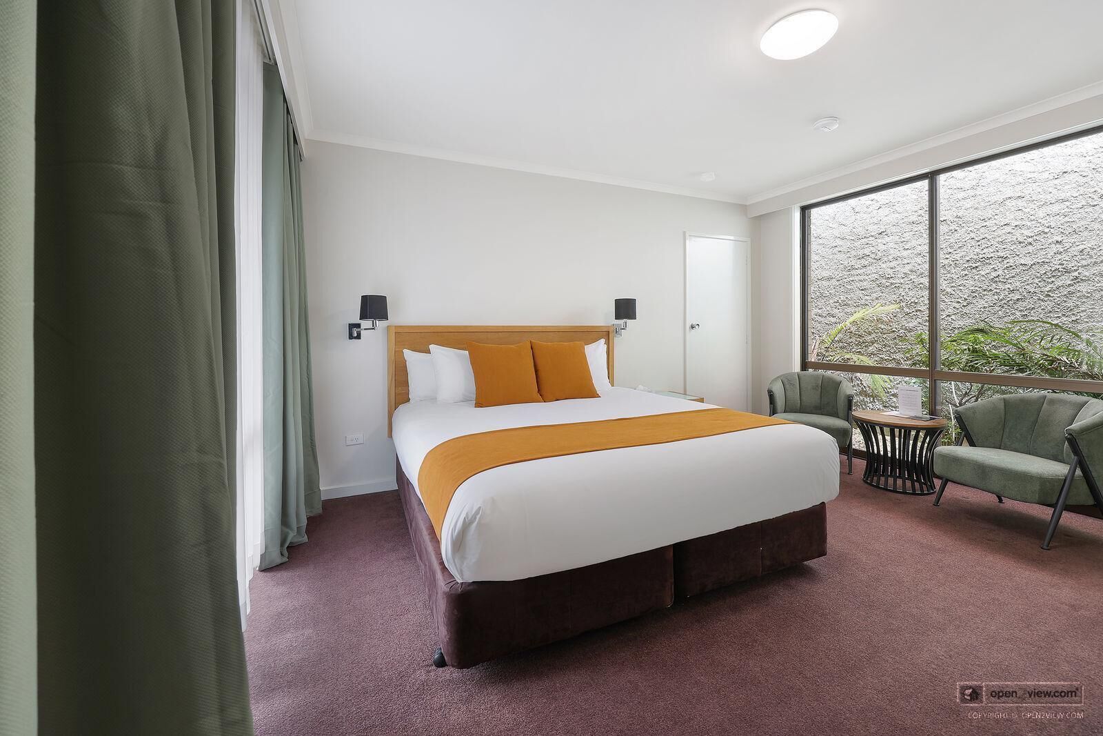 Habitación de hotel con una cama grande, un sillón verde, una mesita y un amplio ventanal con vistas a zonas verdes.