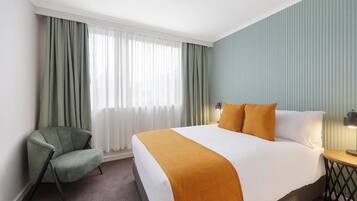 A hotel room with a bed, a chair, a nightstand, and a window with curtains.