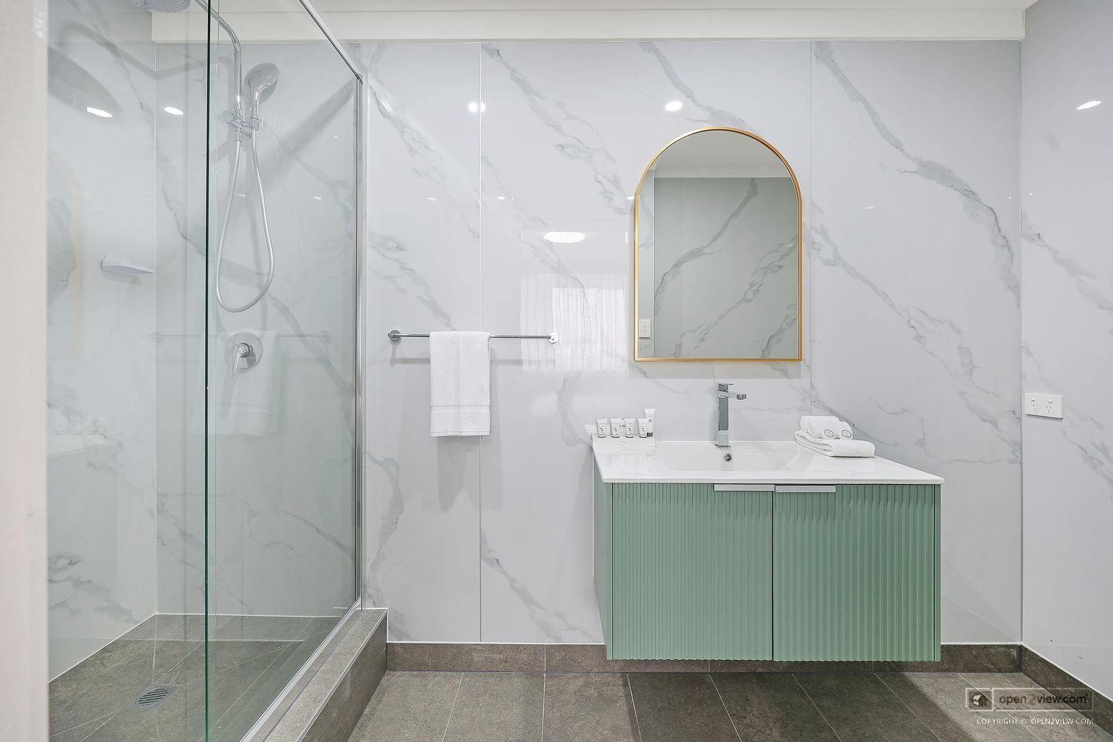 A modern bathroom with a glass-enclosed shower, a marble-tiled wall, a green vanity, and a large mirror with a gold frame.