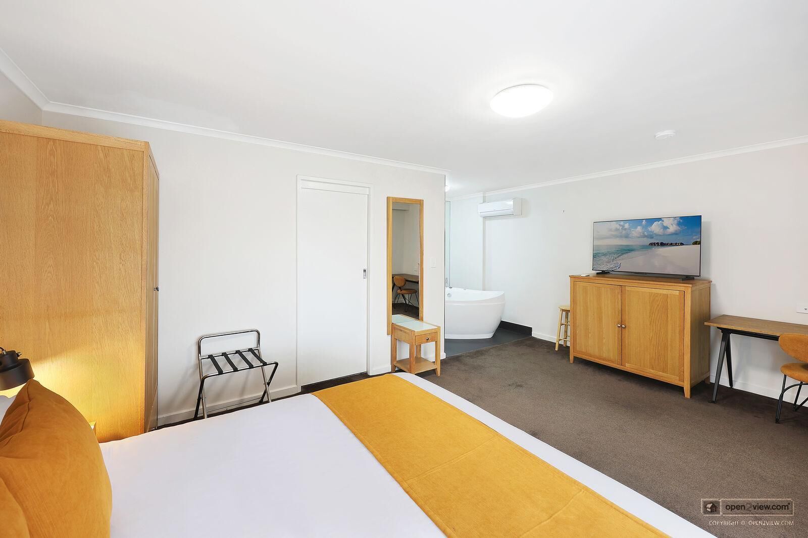 A hotel room with a bed, a wooden wardrobe, a desk, a television, and a bathroom visible through an open door.