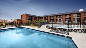 Una piscina con borde de piedra, un edificio con varios balcones y un cielo azul despejado.
