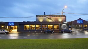 Un motel con un letrero grande que dice "Mid City Motel" y un letrero de "Disponibilidad". Hay coches estacionados adelante, y el edificio tiene un diseño moderno con ventanas grandes.