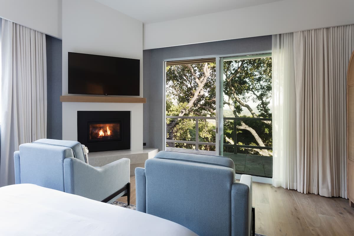 A modern hotel room with a fireplace, a large window with a view of trees, and two blue armchairs.