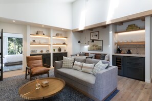 A modern living room with a grey sofa, brown leather chair, and a round coffee table. There are built-in shelves with decorative items and a kitchen area with a sink and cabinets.