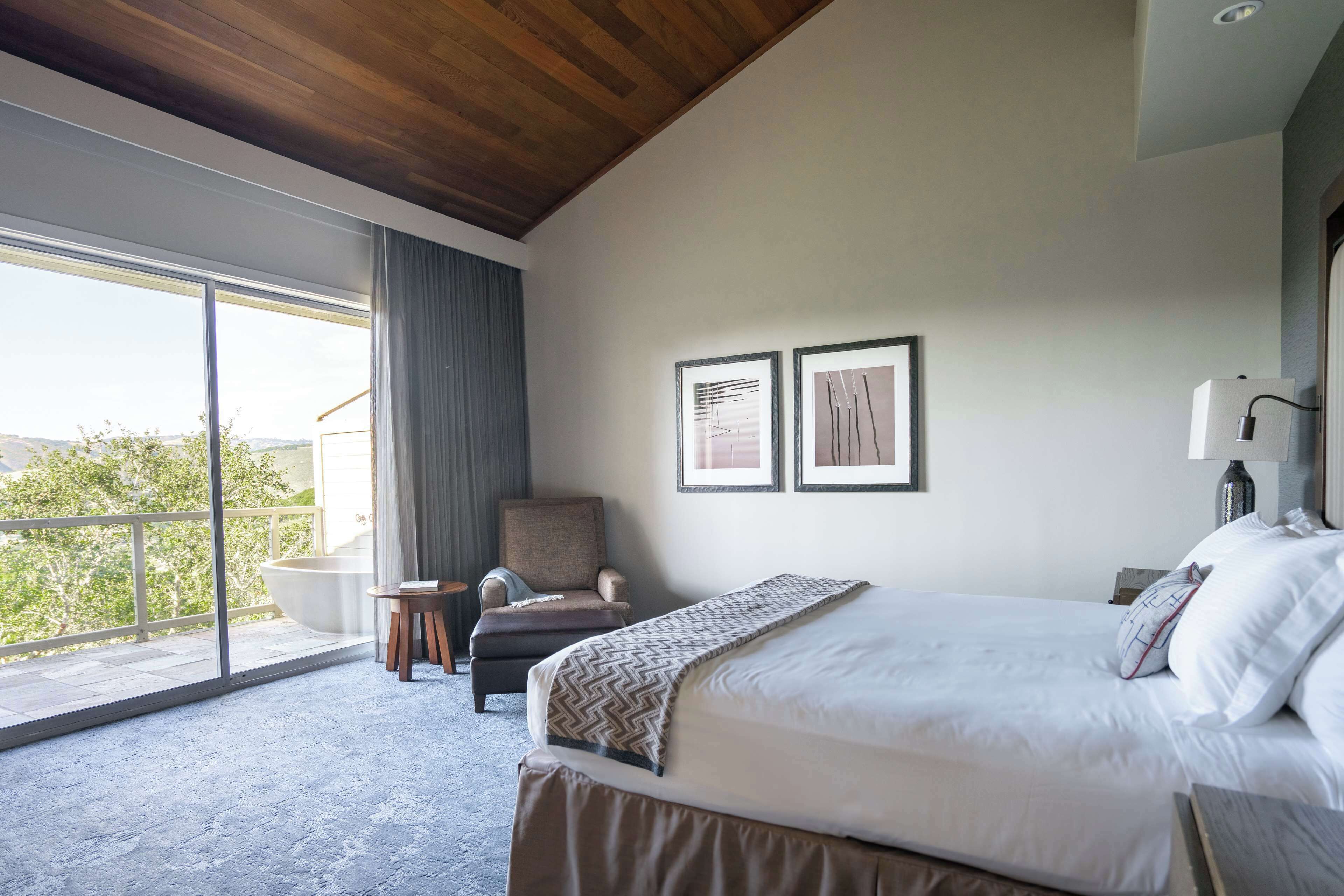 A modern hotel room with a large bed, a chair, a small table, and a view of the outdoors.