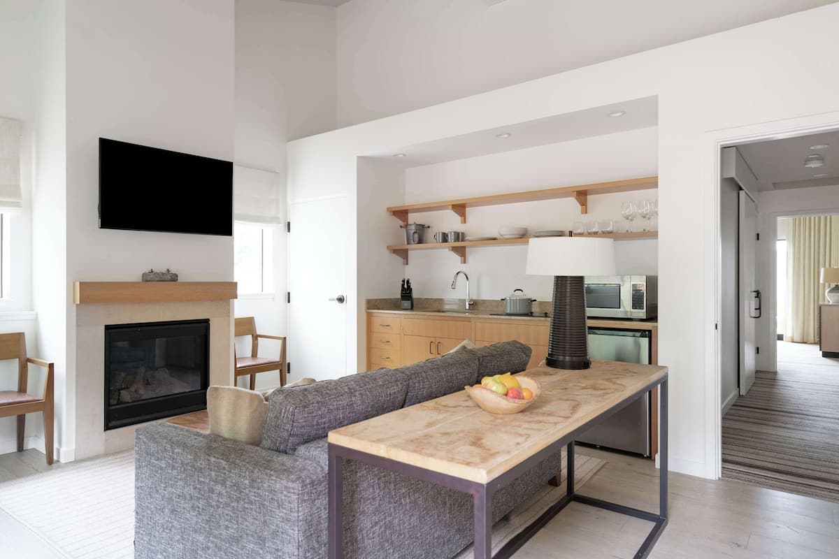 A modern living room with a fireplace, a sofa, a dining table with fruit, and a kitchen area with wooden shelves.